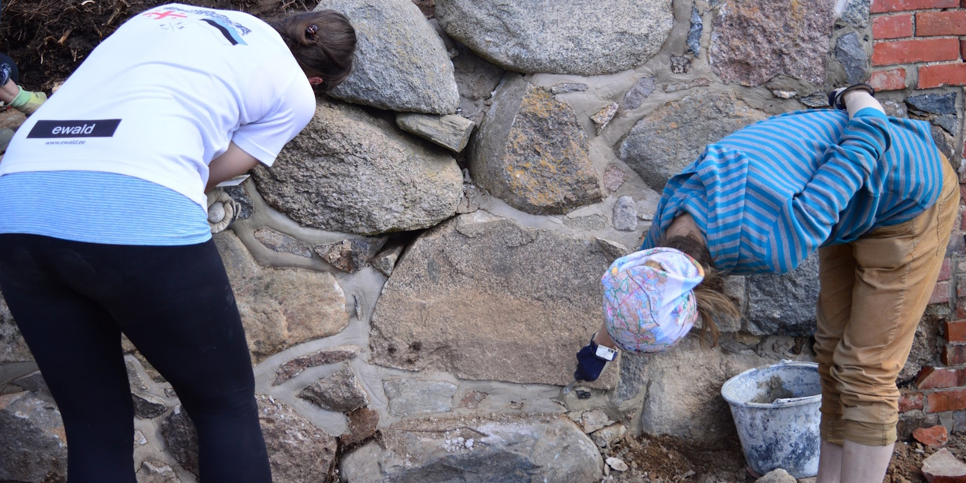 kaks naist on kummardunud maakivimüüri ääres ja parandavad vuugikohti