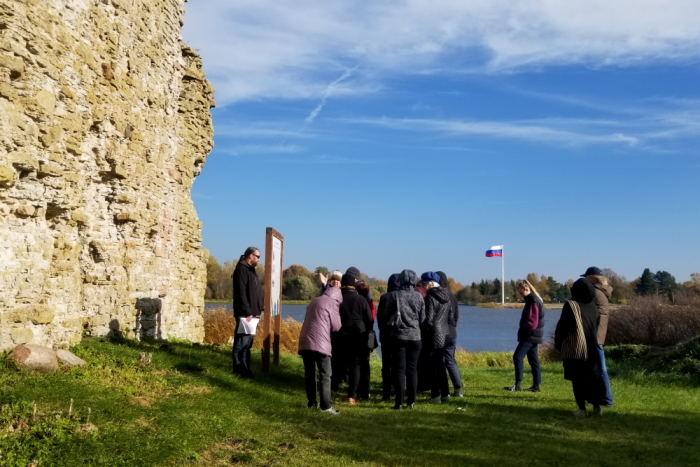 Rühm inimesi seisavad vanade kivimüüride juures. Kauguses näha veeriba ning selle teisel kaldal Venemaa riigilipp.